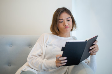 Wall Mural - Skeptical young woman reading book on sofa at home. Pretty Asian lady sitting and studying. Education or literature concept. Front view.