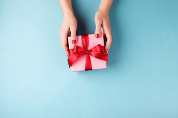 Wall Mural - Overhead view on female hands with red manicure holding pink gift box  with red bow on blue background. Minimal styled composition.