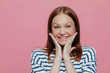 © VK Studio - Headshot of attractive young woman touches cheeks with both palms, has sincere smile, wears casual white and blue striped sweater, isolated over pink background with free space for your text