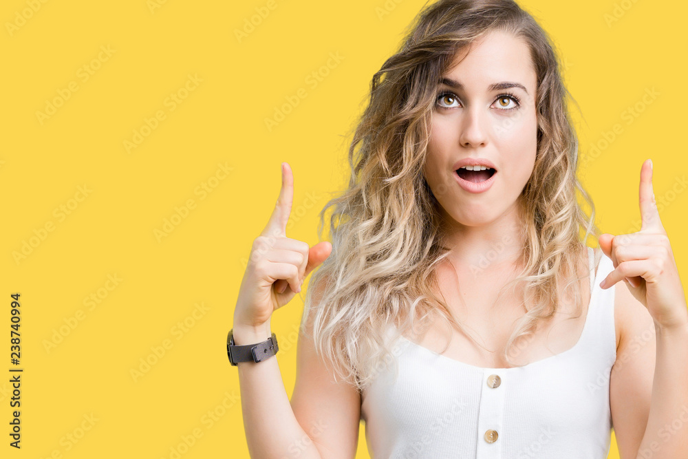 Young beautiful blonde woman over isolated background amazed and surprised looking up and pointing with fingers and raised arms. - obrazy, fototapety, plakaty 