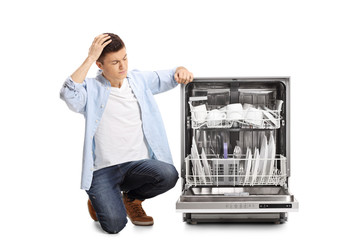 Wall Mural - Angry young guy kneeling next to a loaded dishwasher holding his head