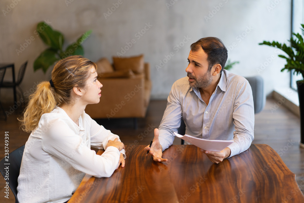 Intern and mentor discussing job specific. Latin man in shirt explaining paper details to young blonde colleague. Mentorship concept - obrazy, fototapety, plakaty 