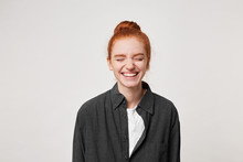 Cheerful Resilient Red-haired Girl With A Bun At Her Head Laughs Sincerely, Having Closed Her Eyes, Dressed In A Simple Black Shirt, Is Isolated On White Background