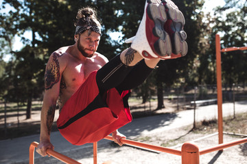 Wall Mural - Young man in headband with naked torso dressed in black leggings and red shorts doing press exercises on the horizontal bar on the sports ground outside on a sunny day