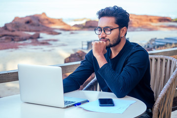 freelancing and remote work.handsome male student working laptop keyboard sitting in cafe on the beach with free internet.Young indian man using computer in summer vacation by the sea