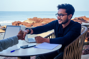 handsome and successful indian man in a stylish well-dressed freelancer working with a laptop on the beach.freelance and remote work.businessman student in a summer cafe on the shore of india ocean