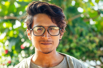 handsome and young indian male model beard and black hair haircut,fashion earring and glasses on a background of green tropical trees.portrait man businessman student relaxing on the beach by the sea