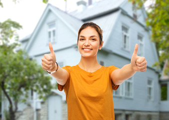 Wall Mural - real estate, gesture and people concept - happy smiling young woman or teenage girl in orange t-shirt showing thumbs up over house background