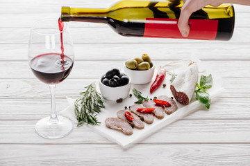 Wall Mural - partial view of woman pouring red wine into glass on white wooden tabletop with meat appetizers near by