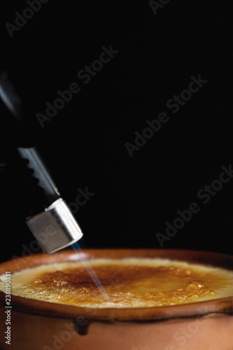 Fotografiet Burning sugar on a caramel custard surface with a home blowtorch in a clay pot with black background