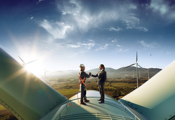 Happy engineer and businessman shake hands after good work. They standing a top of windmill. Around wind generators and beautiful sunset landscape