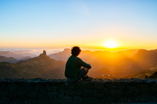 Spain, Canary Islands, Gran Canaria, Back View Of Man Sitting On A Wall Watching Sunset Over Mountainscape