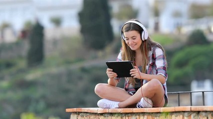 Wall Mural - Happy teen listening and watching media content on a tablet sitting on a ledge on vacation