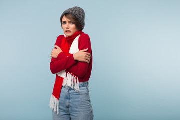 Young unhealthy woman with dark short hair in red sweater,scarf and knitted hat  shaking from cold while sadly looking aside over blue background