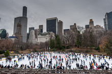 Many People Enjoying Themselves On A Skating Ring In Southern Central Park,  With The Manhattan Skyline In The Background.