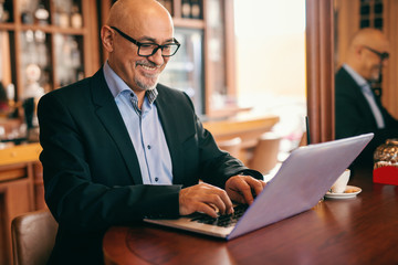 Senior bearded businessman in suit using laptop for work while sitting in cafeteria. On desk coffee.