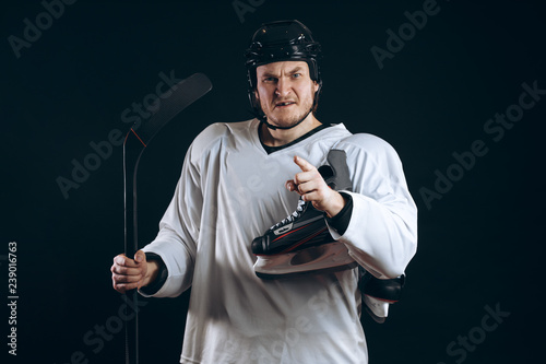 Fototapeta na szkło Portret profesjonalny hokeista w ochronnej odzieży sportowej i hełmie pozuje z kijem hokejowym i jedną łyżwą w rękach na czarnym tle.