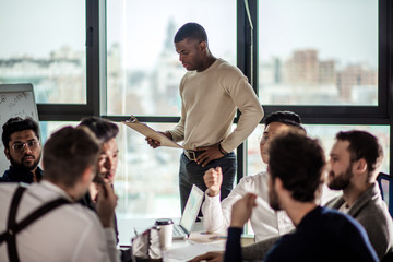 Canvas Print - deadline, technology and people concept - creative team of multiracial men, sitting round the desk in diverse casual and formal clothes talking over new project in office meeting room