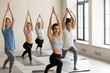 Five multiracial young attractive slim women wearing sportswear standing barefoot on rubber mats in cozy gym studio practising yoga doing warrior pose exercise, improves focus, balance and stability