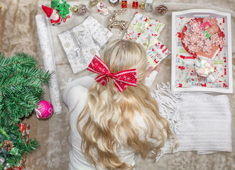 A beautiful young blond woman, with a bow on her head, lies on the floor and wraps Christmas gifts. Christmas preparations. Decorations, Christmas tree, cookies.