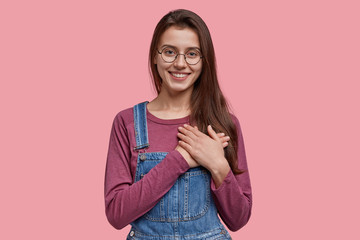 Wall Mural - Kind European woman with pleasant smile, express favour, keeps both hands on chest, being kind hearted and honest, dressed in denim overalls, round optical glasses, isolated over pink background