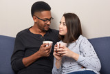 Foto de Horizontal shot of lovely multiethnic girlfriend and boyfriend  drink coffee or tea together, pose at comfortable couch, look at each other  with love, enjoy comfort, togetherness. Interracial relations do Stock