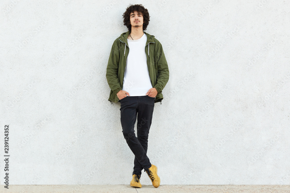 Full length shot of attractive self confident curly man dressed in green anorak, keeps both hands in pockets, enjoys spare time, isolated over white studio wall. People, youth and fashion concept - obrazy, fototapety, plakaty 