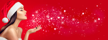 Christmas Winter Fashion Girl On Red Background. Beautiful New Year And Xmas Holiday Makeup. Beauty Model Woman In Santa's Hat Blowing Snow In Her Hand