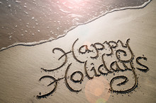 Happy Holidays Message Handwritten In Smooth Sand With An Oncoming Wave In The Lens Flare Of The Tropical Sun