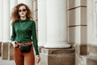 © Victoria Fox - Outdoor fashion portrait of young lady wearing sunglasses, green turtleneck, brown trousers, leather belt bag, necklace, bracelet, posing in street of european city. Copy, empty space for text