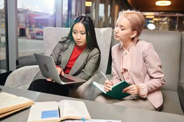 Two young businesswomen sitting at cafe and planning their work together using laptop computer