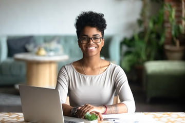 Head shot portrait of happy smiling African American woman sitting at table in cafe, looking at camera, excited female posing, working at computer, doing homework, preparing report in coffee house