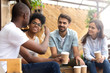 © fizkes - African American man showing thumb up, telling story to interested attentive friends, sitting, drinking coffee in cafe together, smiling multiethnic friends discussing, talking, chatting