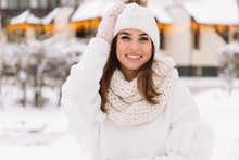 Outdoor Close Up Portrait Of Young Beautiful Happy Smiling Girl Wearing White Knitted Beanie Hat, Scarf And Gloves. Model Posing In Park With Christmas Lights. Winter Holidays Concept.