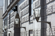 Stylish Iron And Glass Outdoor Wall Lights On The Facade Of A Gray Building Illuminated By Sunlight.
