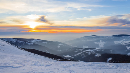 Winter sunrise over Krkonose (Giant Mountains) viewed from the hillside of the Lucni hora, snowy landscape partially covered with fog and colorful cloudy sky with sun shining through the clouds
