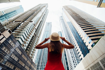 Back view of young woman in red summer dress holding with both hands straw hat looking up on skycrapers at downtown in modern city. Low angle view.