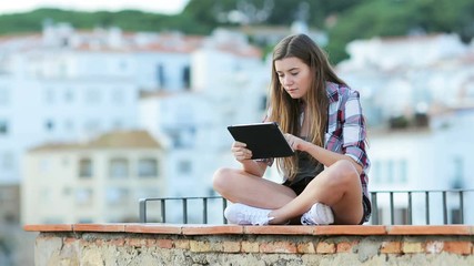 Wall Mural - Serious teen using a tablet on vacation sitting on a ledge in a town