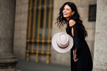 beautiful happy young girl in a black dress and hat is resting outdoors, amazing woman is walking in an urban environment