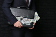 © Pixel-Shot - Businessman holding briefcase with dollar banknotes on dark background. Corruption concept