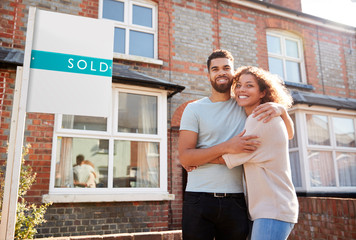 Portrait Of Excited Couple Standing Outside New Home With Sold Sign