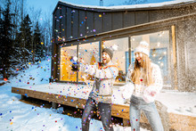 Young Happy Couple Dressed In Sweaters Celebrating Winter Holidays In Front Of A Beautiful Decorated House In The Mountains