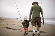 Young Boy And His Father Walking Along A Beach Carrying Fishing Rods And Wearing Hats.