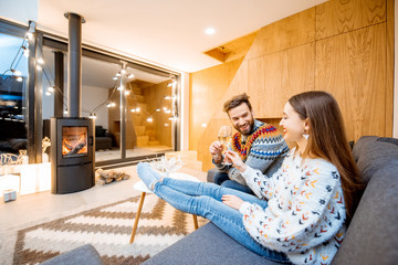 Young couple celebrating winter holidays sitting with wine on the couch in the beautiful decorated house with fireplace