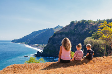 Wall Mural - Family vacation lifestyle. Happy mother, kids on hill with scenic view of high cliffs, fishers village on black beach. Children looking at blue sea. Bukit Asah is popular travel destination in Bali.