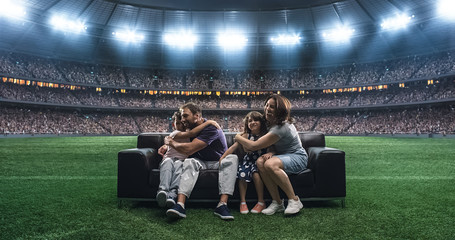 Wall Mural - A family is watching a soccer moment, sitting on the couch located in the middle of the soccer stadium.