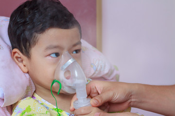 Close-up nurse’s hand helping sick child hold nasal mask with respiratory problem in hospital room. Asian boy patient inhalation therapy by the mask of inhaler with bronchodilator’s soft stream smoke