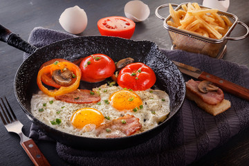 Fried eggs with bacon and tomatoes on an old cast-iron pan with french fries on a gray table. Close-up