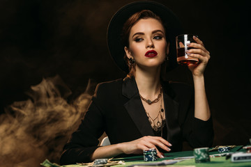 attractive girl in jacket and hat holding glass of whiskey at poker table in casino and looking away