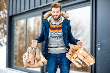 Handsome man in winter clothes carrying firewoods on the terrace near the modern house in the mountains